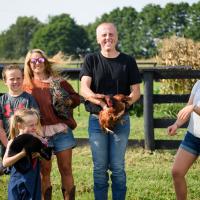 Dr. Day stands in the middle of the photo, holding a chicken that is trying to fly away. His wife and four children stand around him, holding cats and a family dog. Dr. Day is smiling despite the chaos around him.