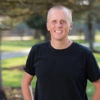 A portrait photo of Dr. Day smiling with greenery in the background. He is wearing a plain black t-shirt and is standing with his hands on his hips.