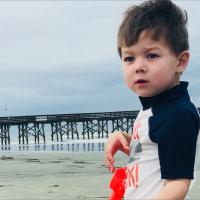 Marco stands on the beach in with a pier in the distance behind him.