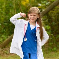 Taytum, wearing blue scrubs, a white doctor’s coat and a pink stethoscope, flexes one arm and smiles.