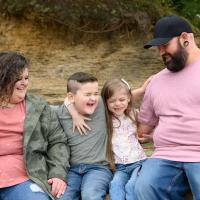 Taytum and her family all laugh as they hug each other. They are sitting on some rocks in an outdoor setting.