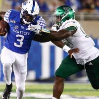 An action shot of Terry running with a football tucked under his right arm as his left pushes away the opposing team’s player who is grabbing his left shoulder.