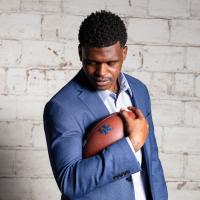 A portrait of Terry, dressed in a blue blazer and light blue button-up shirt, cradling a football with his right arm while looking down.