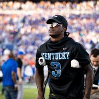 Terry, dressed in a short-sleeved black hoodie, hat and sunglasses, stands on crutches with a blurred background of fans in the stadium seats behind him.