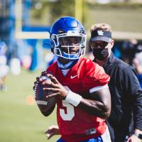 Terry, dressed in practice gear and in a red jersey to signify him as the quarterback, during a practice with a coach and fellow teammates in the background behind him.