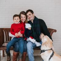 Dr. London-Bounds is seated with her two kids, a young brown-haired boy with a red sweater and jeans, and a baby girl in white tights, red dress and fur navy winter jacket. Dr. London’s husband, a middle-aged white man in a black sweater with a dark blue and green checkered scarf, sits next to her. They are seated on a couch outside, as their golden labrador retriever lays down beside them.