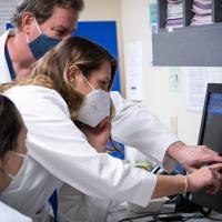 Dr. London-Bounds is dressed in her white doctor’s coat and mask, standing in a medical office between two of her colleagues dressed in the same gear—an older white man with grey hair, and a younger whihte woman with brown hair. Dr. London is pointing at the screen at the same time as her male colleague, while simultaneously being on the phone.