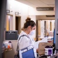 Dr. London-Bounds is standing in the hallway of UK HealthCare reading through notes. She is wearing her doctor’s coat and mask, and has her hair tied into a bun.