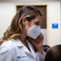 Dr. London-Bounds stands with her phone to her ear in a medical office. She is wearing her doctor’s coat and a mask.