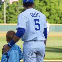 T.J. stands next to a young kid with short hair and a tie-dye shirt. The two are standing on the edge of a baseball field, watching other players warm up. T.J. has his hand on the kid’s shoulder.