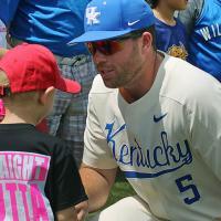 T.J. crouches down to talk to two kids. He has sunglasses and a baseball uniform on and there is a line of fans and baseball players behind him.