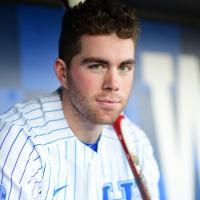 A portrait of T.J., with his face in focus and the background blurred. He is sitting in the dugout and has a baseball bat over his shoulder. He is wearing his baseball uniform and has a slight smile on his face.