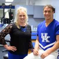 Travis smiles for the camera alongside Dr. Kimberly Kaiser, a white woman with long light-blonde hair. She is wearing a black, long-sleeve shirt with lace detailing and a large blue gemstone necklace.