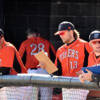 Trent is reading off a clipboard, standing next to his teammates on a baseball field in his number 13 Tigers baseball uniform, cap and shades.