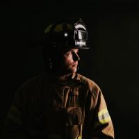 Trent in his firefighting gear and uniform, in front of a black background. His head is turned to the side for a profile view.