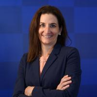 Dr. Ashley Montgomery-Yates, a middle-aged white woman with brown hair, stands smiling inside the Kroger Field stadium in front of a blue checked background. She is wearing a navy suit, a pearl necklace with matching earrings.