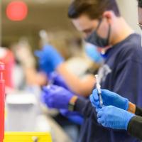 A row of medical staff members, dressed in scrubs, masks and gloves, at the Kroger Field Vaccine Clinic are disassembling syringes in the proper medical manner in order to be disposed of.