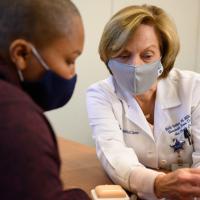 A close-up photo of Dr. Beth Holden, an older white woman with short blonde hair, showing a device Whitley. Dr. Holden is wearing a white lab coat over a white shirt, and a gray face mask.