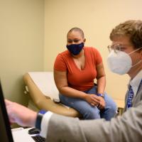 Whitley looks at the computer screen of Dr. Kyle Rosenstein, a white man with wavy light brown hair, who is pointing at the screen. He is wearing a light gray suit jacket over a white button-up shirt, an N-95 face mask, and a pair of light-rimmed glasses.