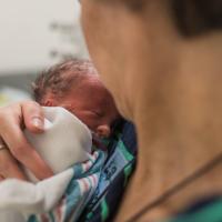 An over-the-shoulder shot of Karla Duerson, a white woman with short blonde hair, holding Wylie shortly after she was born. Karla is wearing a blue and green striped long-sleeve sweater.