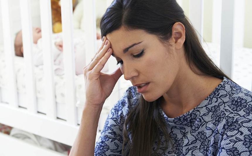 A mother with an infant in a crib rests her forehead on her fingers.